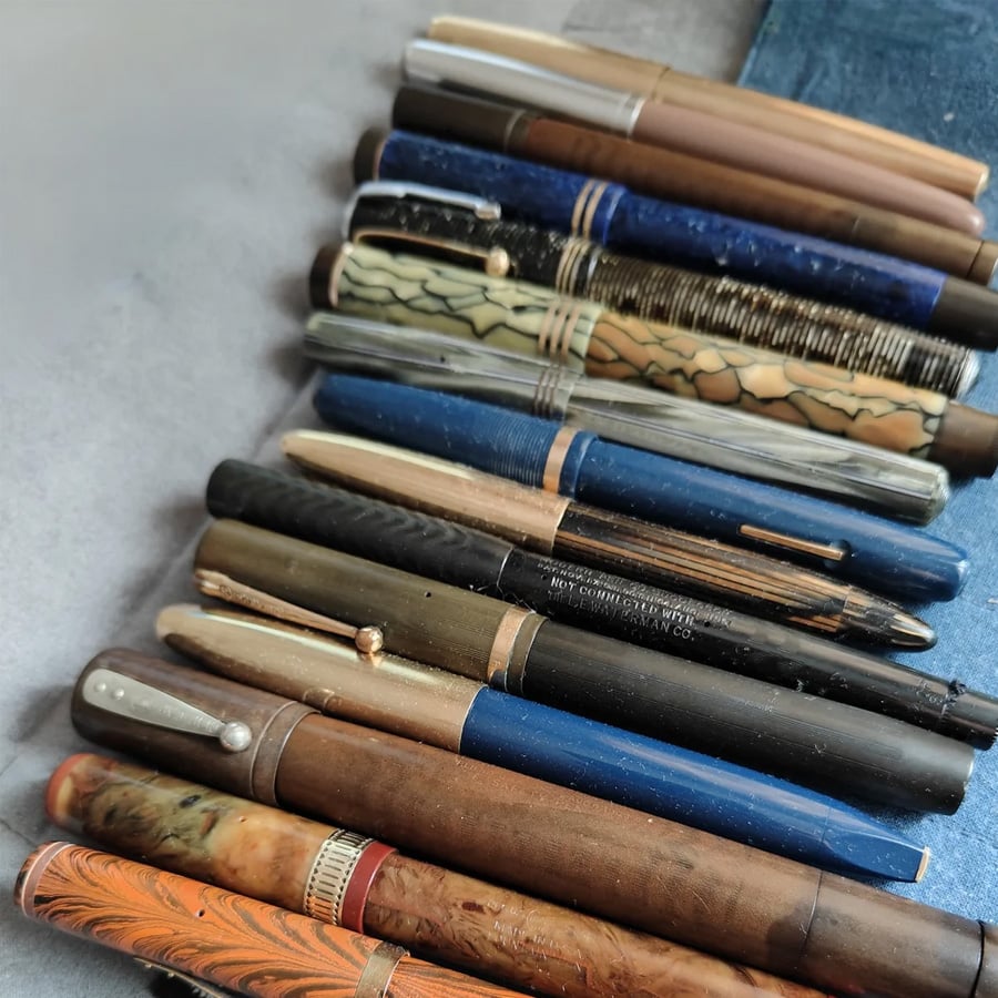 A collection of vintage fountain pens with various colors, patterns, and materials are neatly arranged on a flat surface next to a blue notebook.