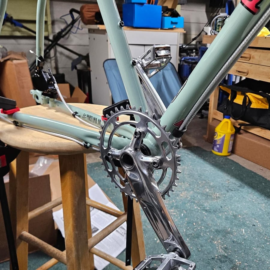 Close-up of a partially assembled light blue bicycle frame on a wooden stool indoors, showing the crankset and pedals, with tools and supplies scattered in the background.
