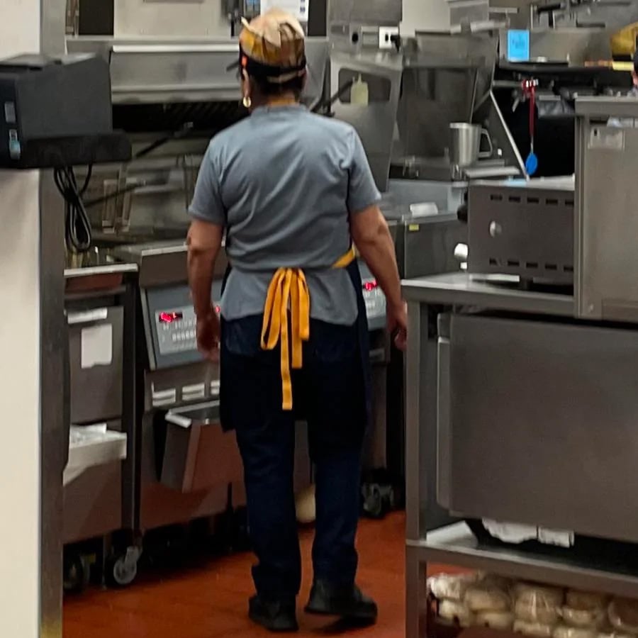A person wearing a gray shirt, dark pants, and a yellow apron stands in a commercial kitchen surrounded by stainless steel equipment and cooking appliances.