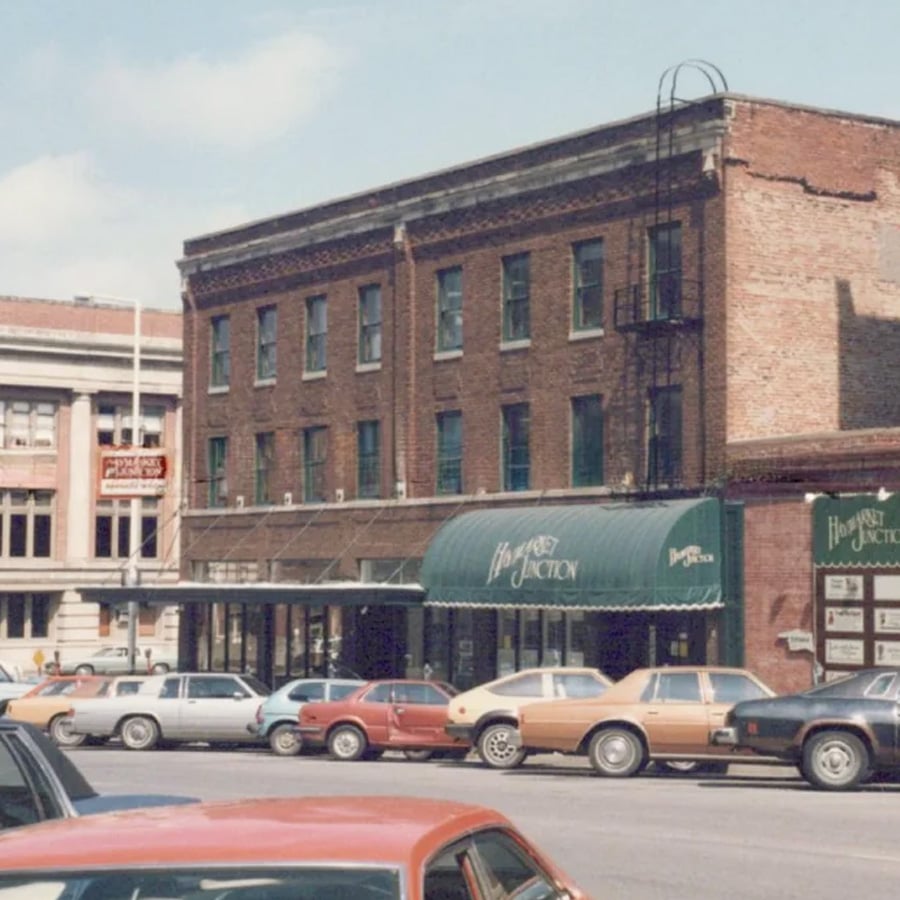 A row of vintage cars is parked in front of a brick building with green awnings labeled “Harvest Junction” on a sunny day. Another building with columns is visible in the background.