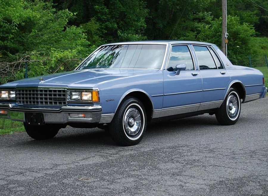 A light blue, four-door classic sedan parked on a paved road, with green trees and grass in the background.