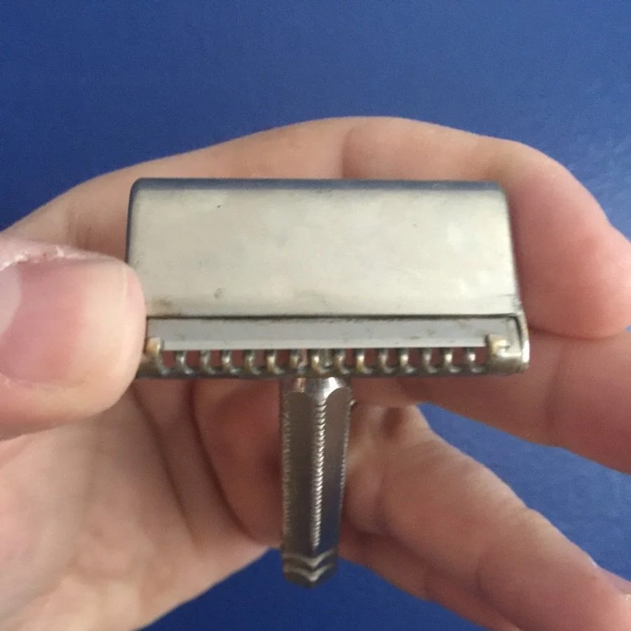 A hand holds a vintage metal safety razor with a textured handle, set against a solid blue background.