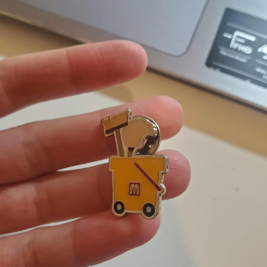 A hand holds a pin shaped like a yellow cleaning cart with a mop and caution sign, featuring the McDonald’s “M” logo.