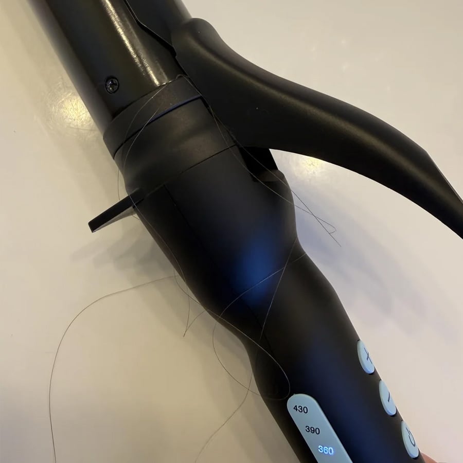 A black curling iron with long strands of hair tangled around the barrel and handle, lying on a white surface.