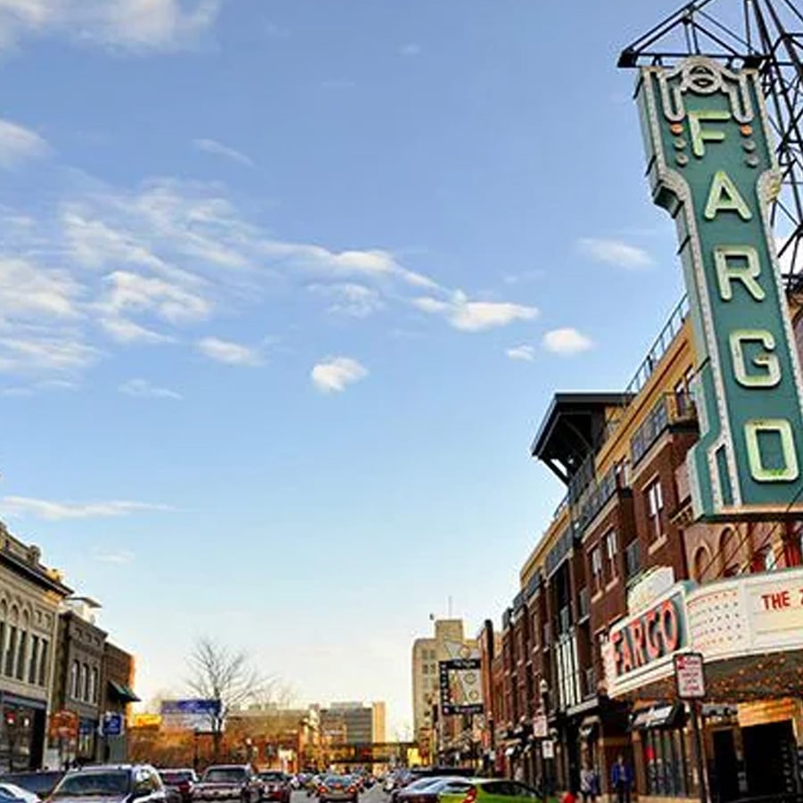 Street scene in downtown Fargo, North Dakota, featuring historic buildings, cars parked along the road, and a large vertical Fargo theater sign against a blue sky with scattered clouds.