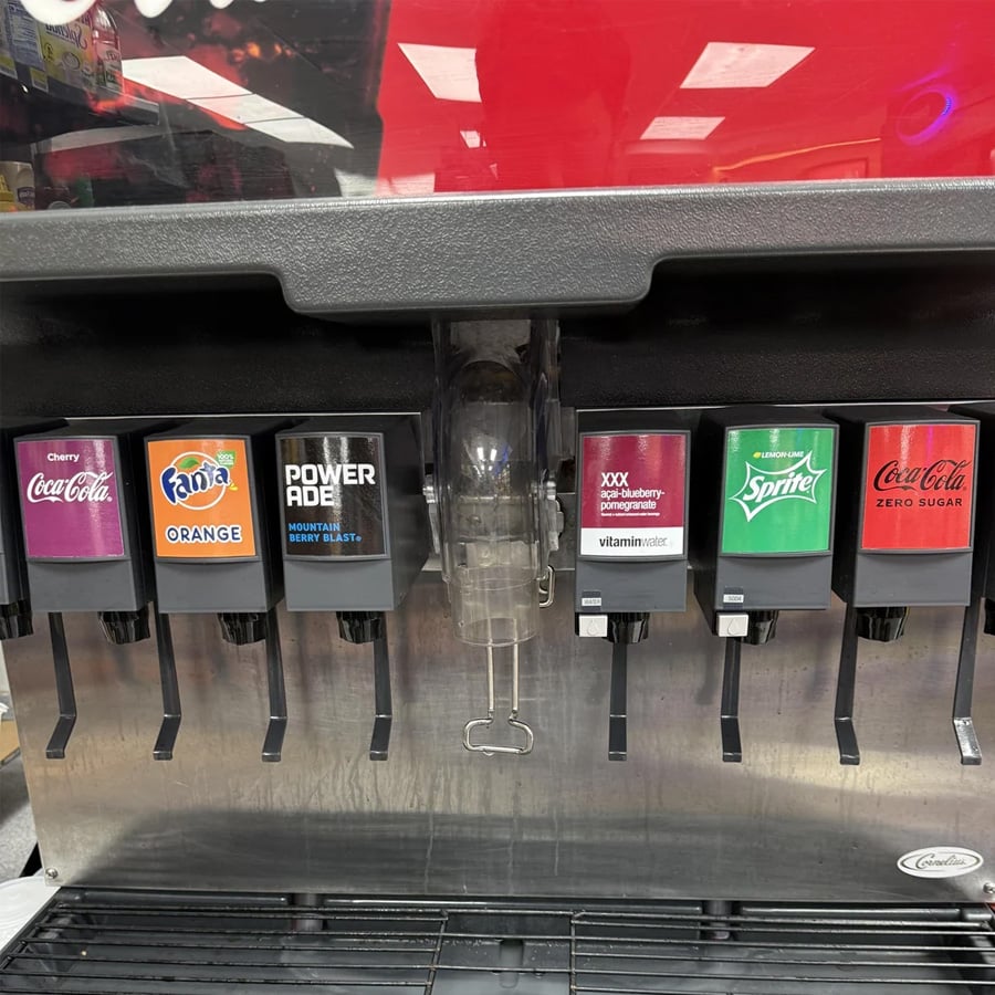 A soda fountain machine with dispensers for Cherry Coke, Fanta Orange, Powerade, Vitaminwater, Minute Maid Lemonade, Sprite, and Coke Zero Sugar. The background shows a red surface.