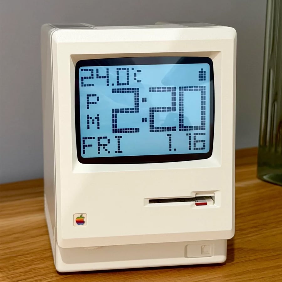 A retro-style digital clock resembling an old Macintosh computer displays the time 2:20 PM, 24.0°C, Friday, and 1.75. It rests on a wooden surface near a glass object.