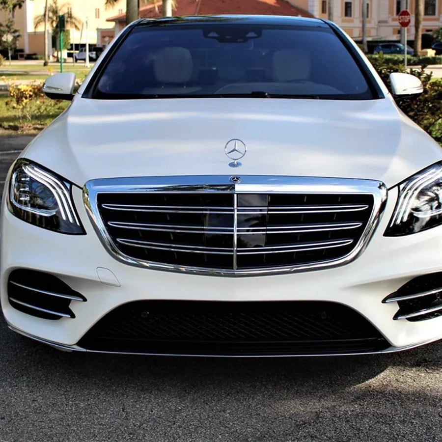 Front view of a white Mercedes-Benz sedan parked on a street, showcasing the chrome grille and emblem, with buildings and palm trees visible in the background.