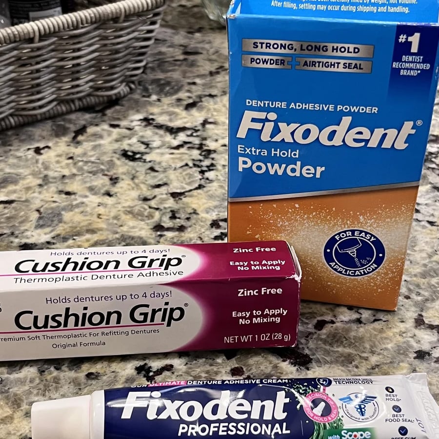Three dental adhesive products—Fixodent Extra Hold Powder, Cushion Grip Thermoplastic Denture Adhesive, and Fixodent Professional—are displayed on a granite countertop with a basket visible in the background.