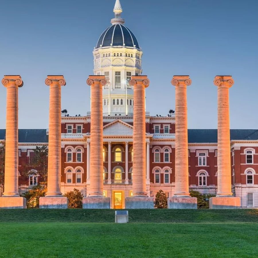 Six stone columns stand on a lawn in front of a historic red brick building with a white dome and clock, under a clear blue sky.