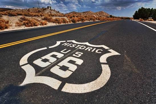Close-up of a Historic Route 66 emblem painted on an empty highway in a desert landscape, with hills, dry shrubs, and a partly cloudy sky in the background.