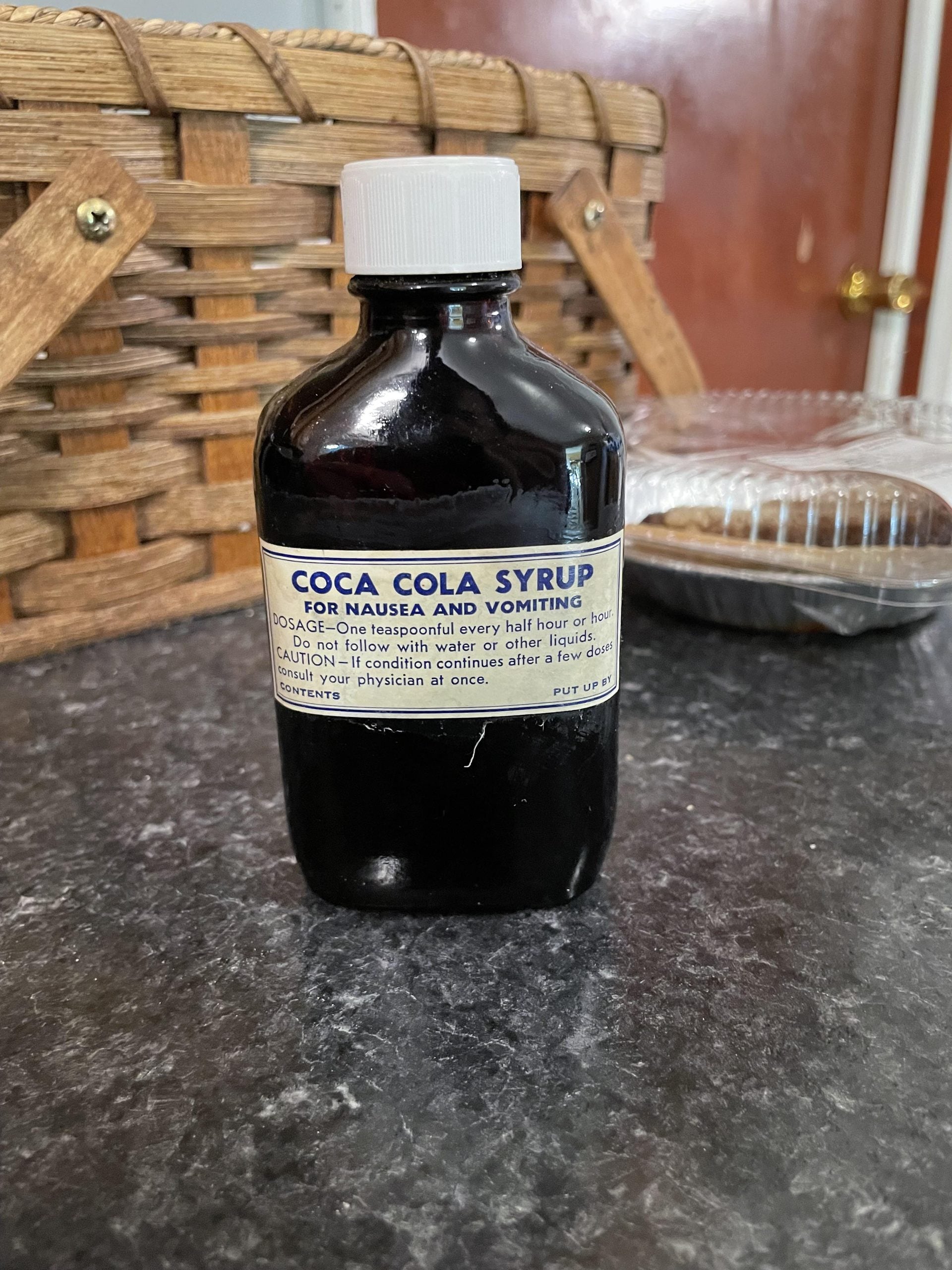 A dark glass bottle labeled “Coca Cola Syrup for Nausea and Vomiting” sits on a gray countertop. A wicker basket and a clear plastic food container are visible in the background.