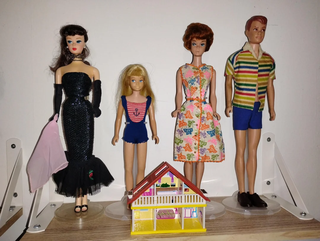 Four vintage dolls stand in a row behind a small toy house. Two female dolls wear dresses, one wears a blue swimsuit, and the male doll wears a striped shirt and blue shorts. All are displayed on a shelf.