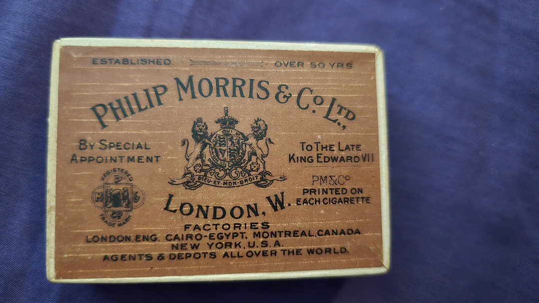 A vintage wooden Philip Morris & Co. Ltd. cigarette box with ornate text and a royal crest, mentioning factories in London, New York, Montreal, and other cities, on a dark blue fabric background.