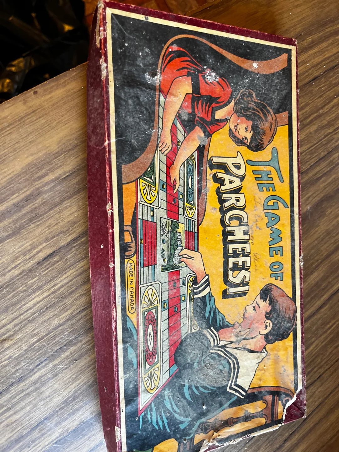A vintage “The Game of Parcheesi” box with colorful artwork showing a girl and boy playing the board game on a wooden surface. The box has visible wear and tear.