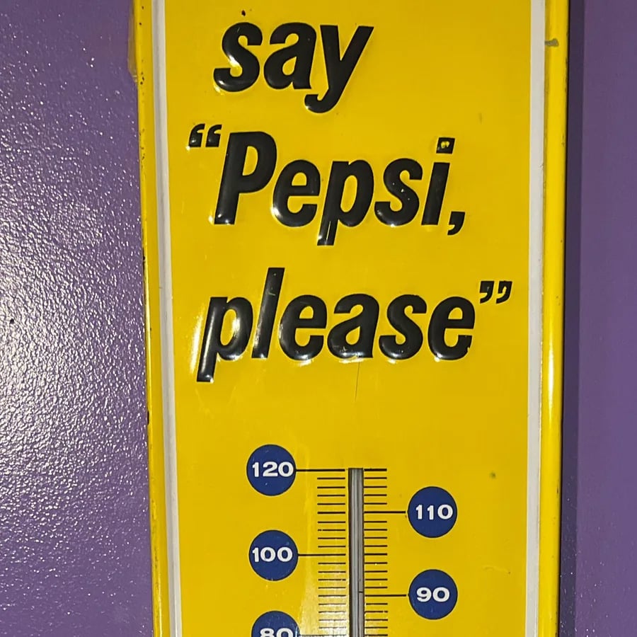 A yellow vintage sign with black text reads ‘say “Pepsi, please”’ and features a thermometer below, against a purple wall.