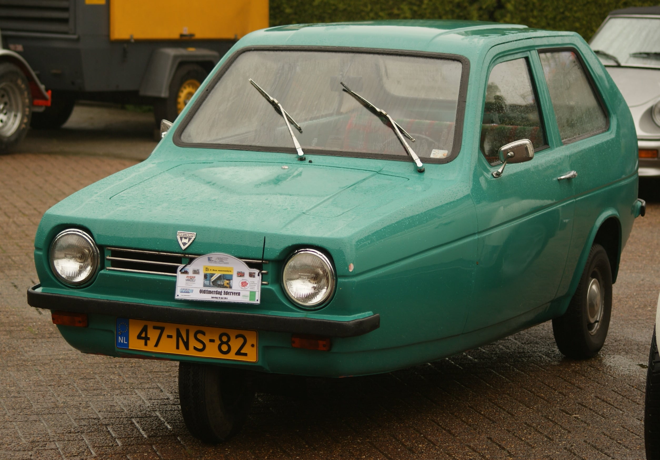 A green three-wheeled car with a Dutch license plate is parked on wet pavement. The car has round headlights, dual windshield wipers, and a sign displayed on the front. Other vehicles are visible in the background.