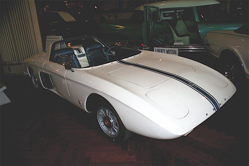 A vintage white convertible sports car with black racing stripes is displayed indoors on a wooden floor. Classic cars are partially visible in the background.