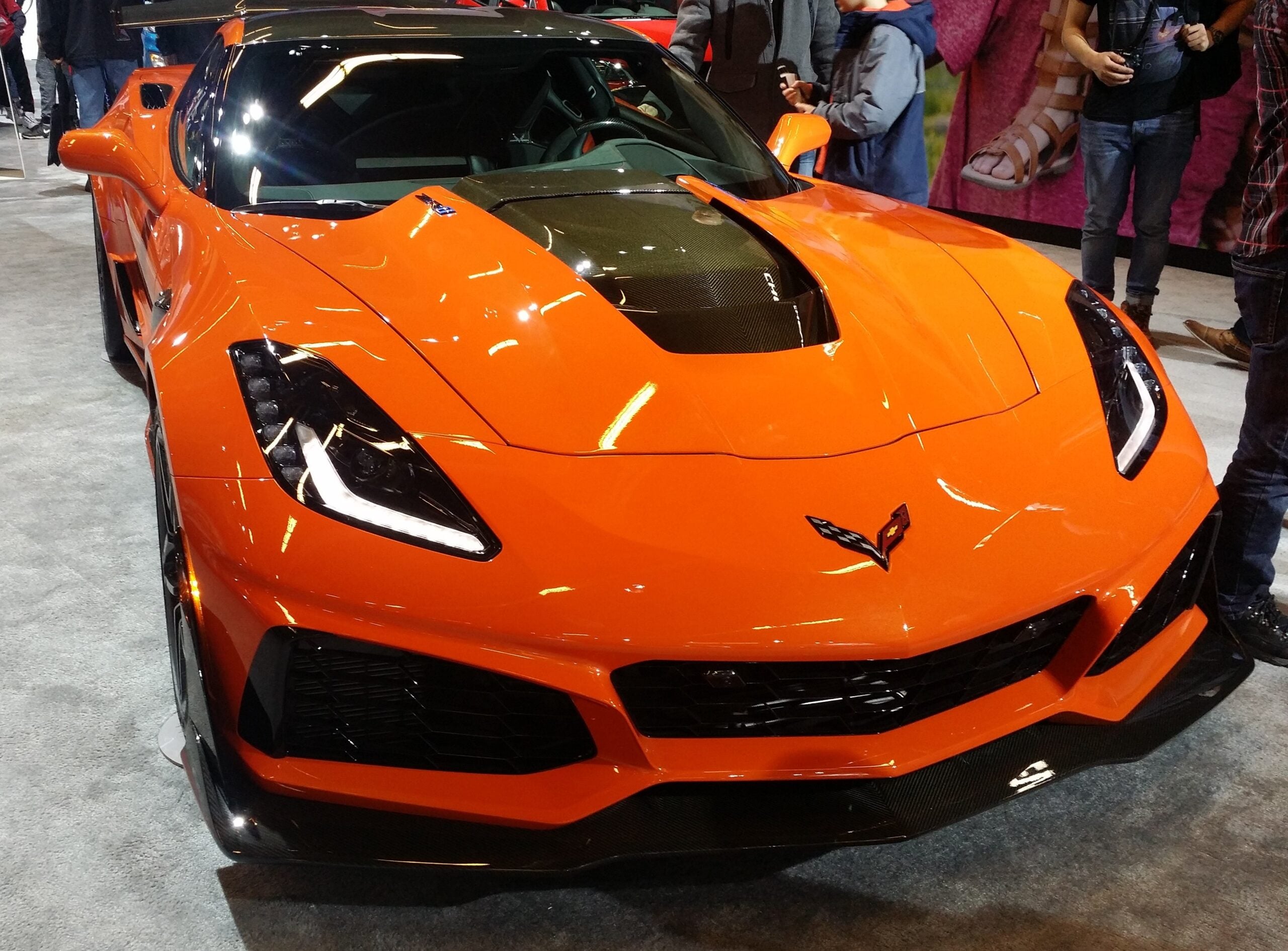 A bright orange Chevrolet Corvette sports car on display at an auto show, surrounded by people. The car features a black hood vent and sharp, angular headlights.