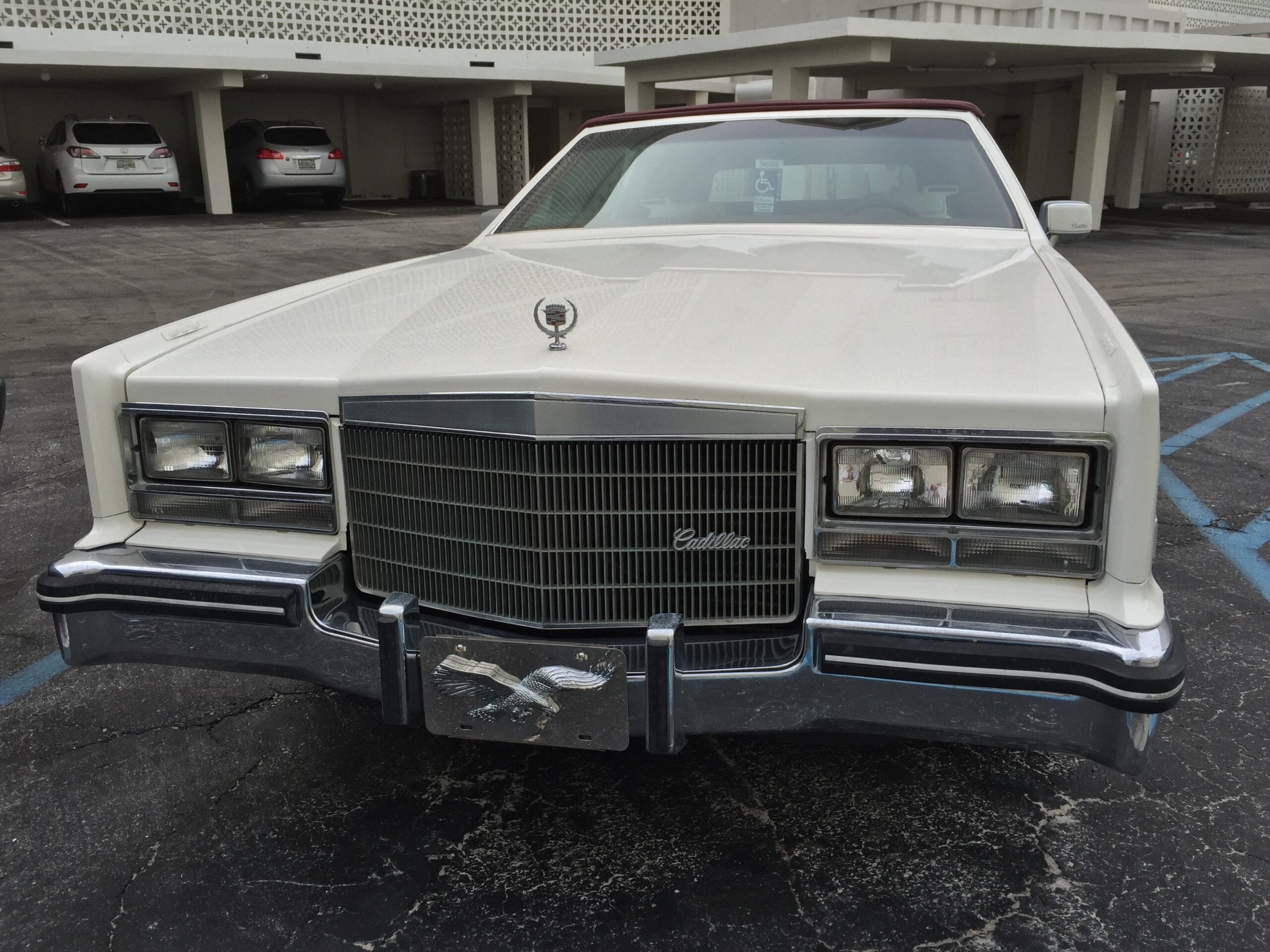 A white vintage Cadillac parked in a lot, viewed from the front, with a large chrome grille and rectangular headlights. The background shows a white, multi-level building and other parked cars.
