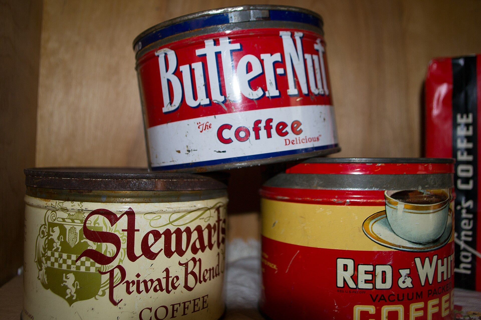 Three vintage coffee tins are stacked together on a shelf. The tins are labeled “Butter-Nut Coffee,” “Stewart’s Private Blend Coffee,” and “Red & White Vacuum Packed Coffee.”