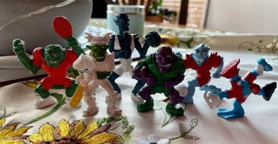 Six colorful monster figurines, each dressed in sports attire and posing as athletes, stand on a table with a floral embroidered cloth and household items in the background.