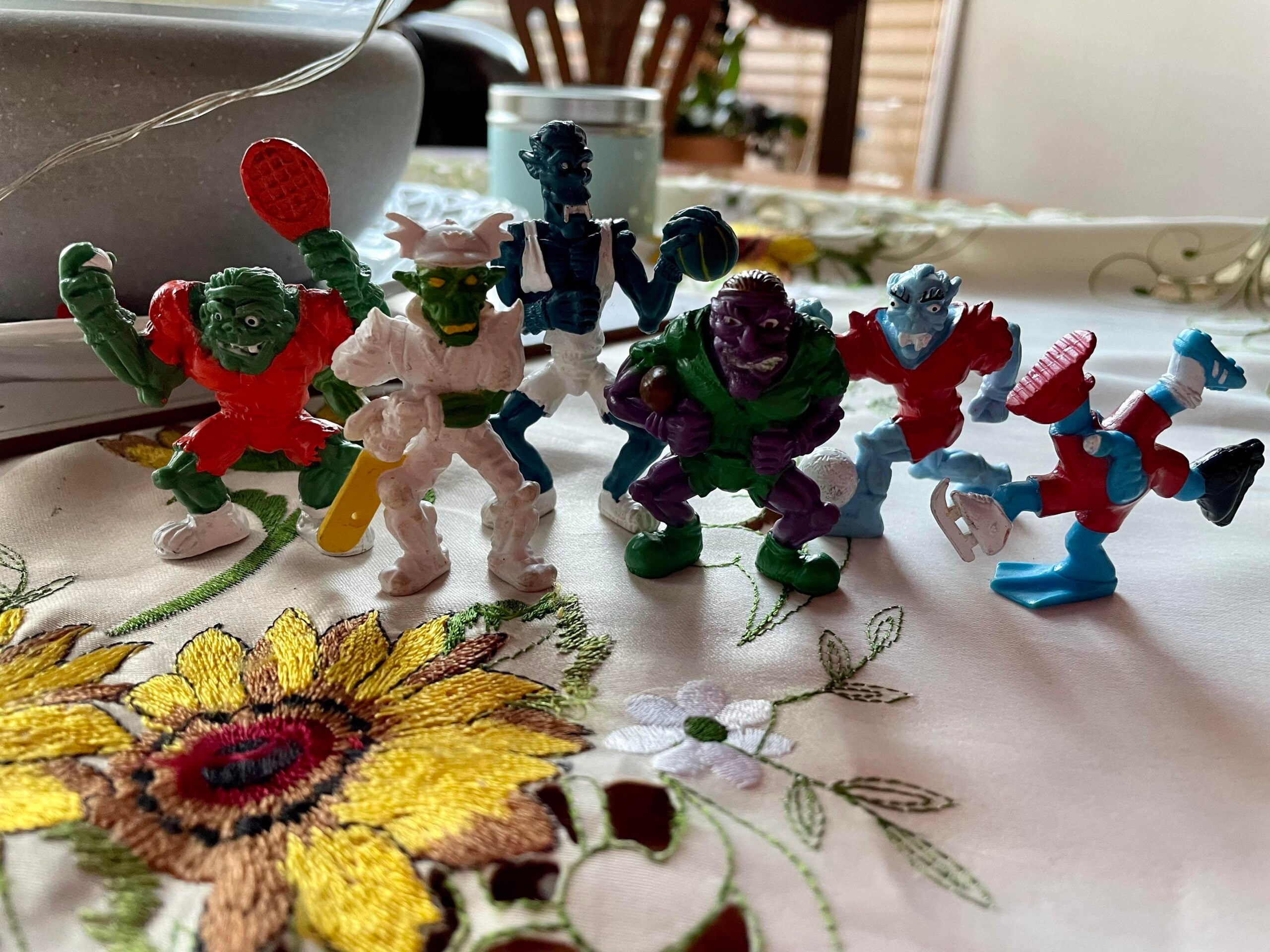 Six colorful monster action figures stand on a tablecloth with flower embroidery, posed in dynamic stances. A gray bowl and potted plant are visible in the background.