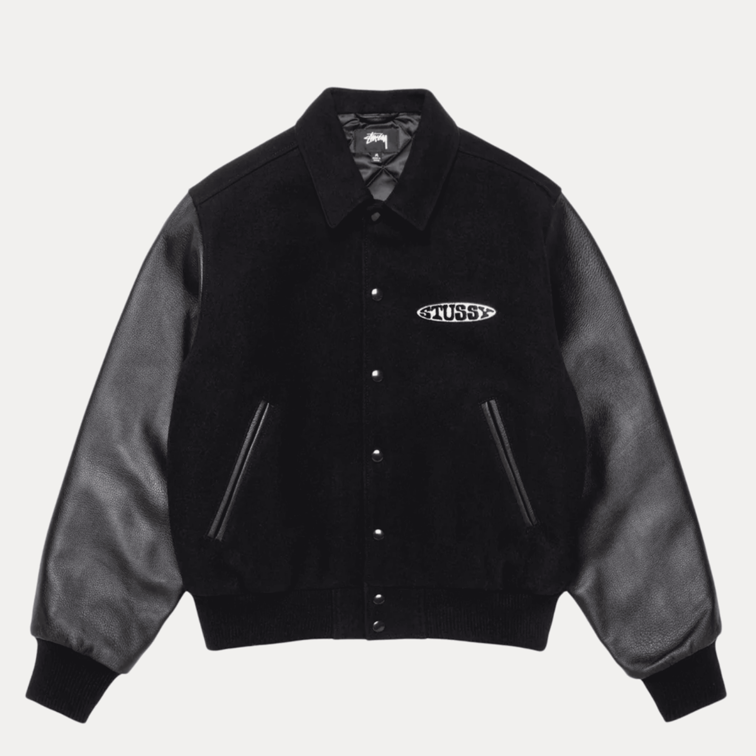 Black varsity jacket with leather sleeves, snap buttons, and “STUSSY” logo patch on the left chest. The jacket has ribbed cuffs and waistband.