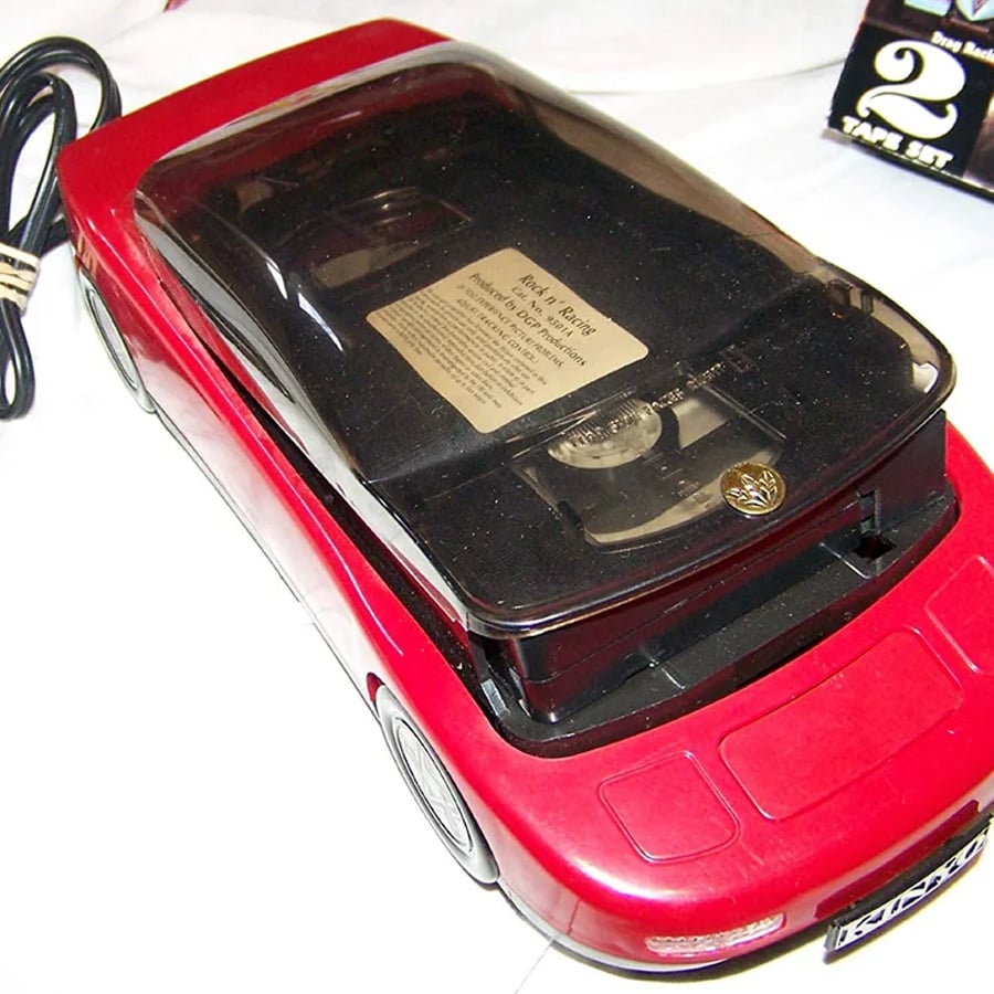 A red car-shaped VHS tape rewinder with a clear black plastic cover showing the tape inside, sitting on a white surface.