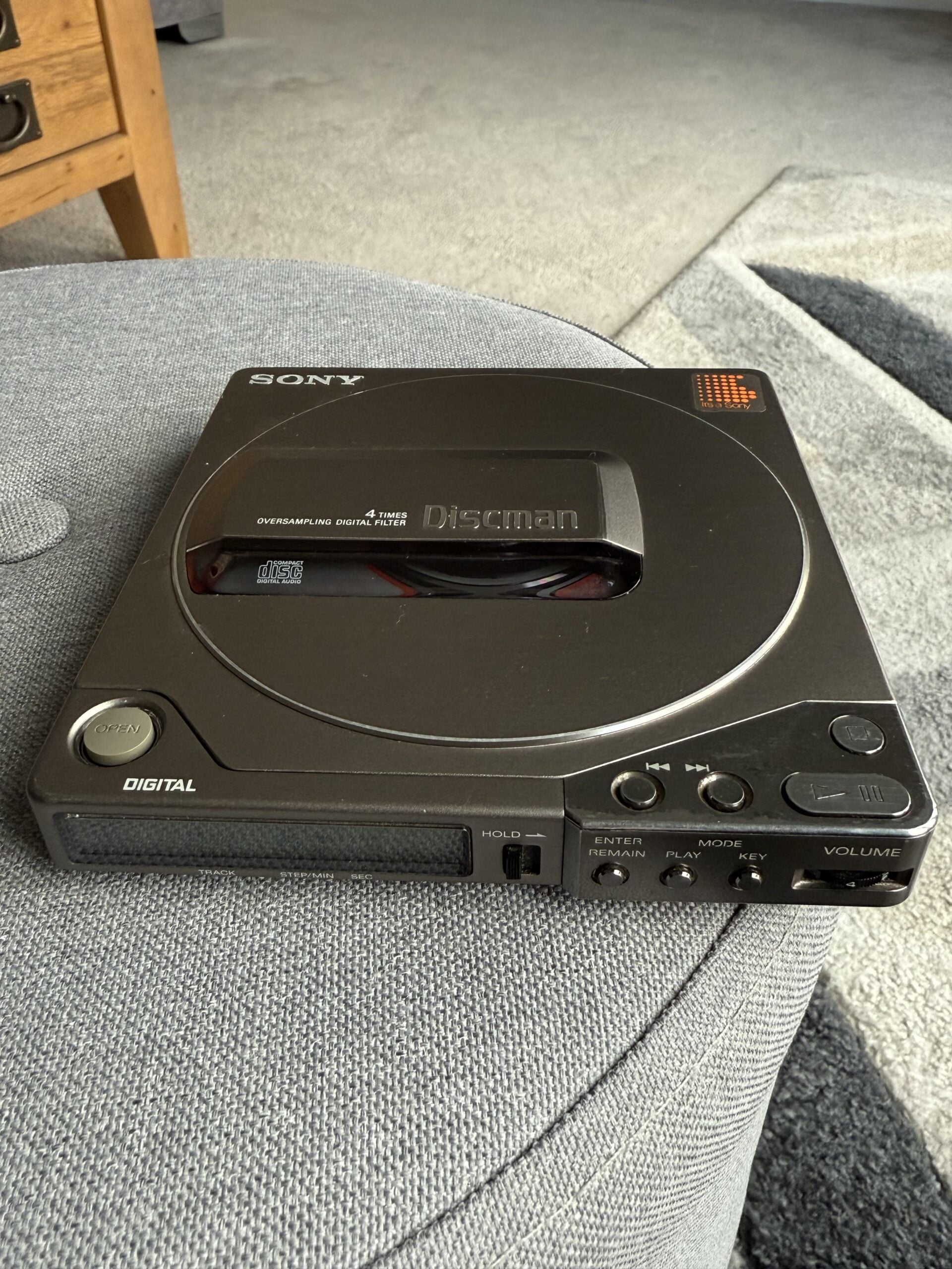 A black Sony Discman portable CD player sits on a light gray fabric surface, with buttons and display visible on the front panel.