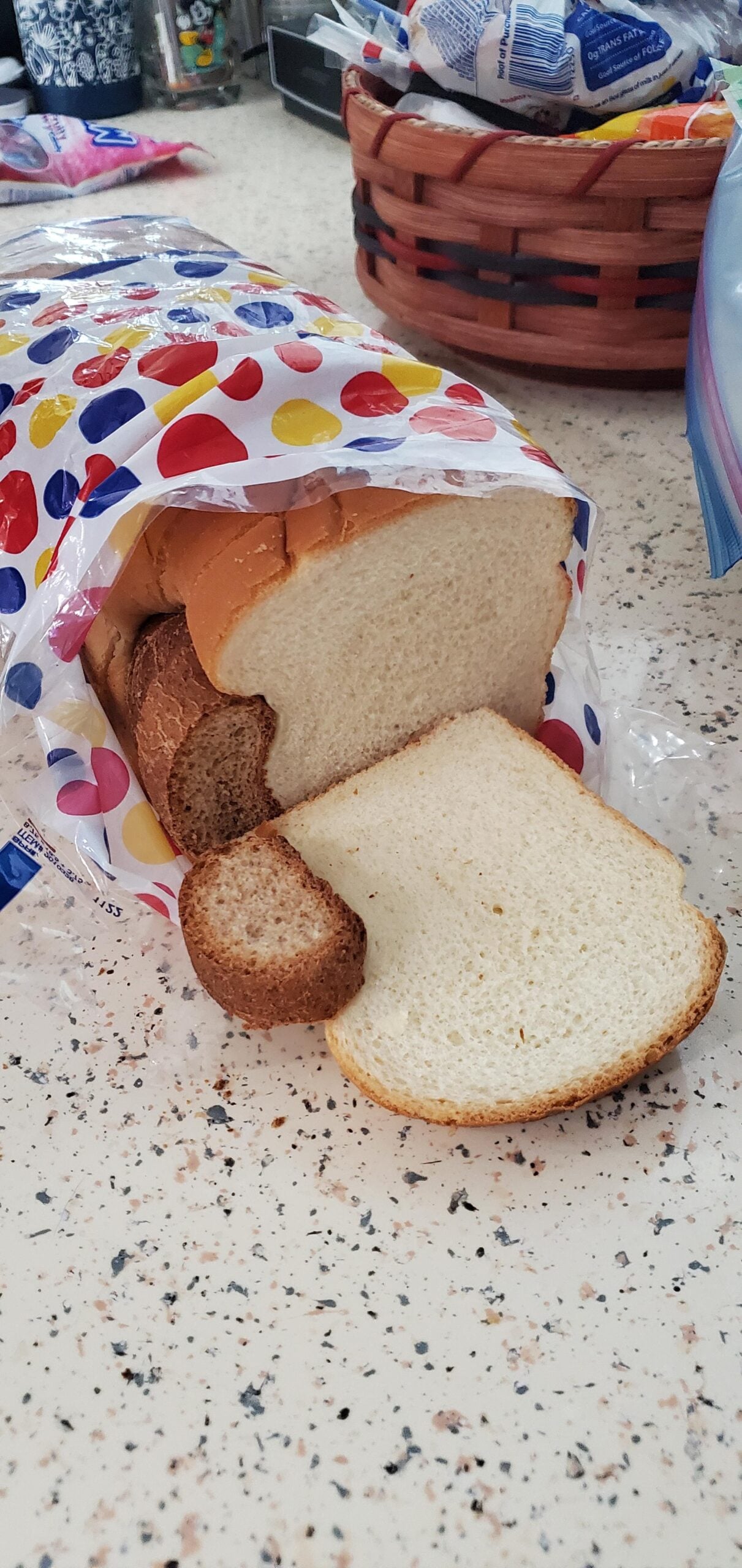 A loaf of sliced white bread in a colorful, dotted plastic bag sits on a speckled kitchen counter, with two brown end pieces and a white slice lying at the front.