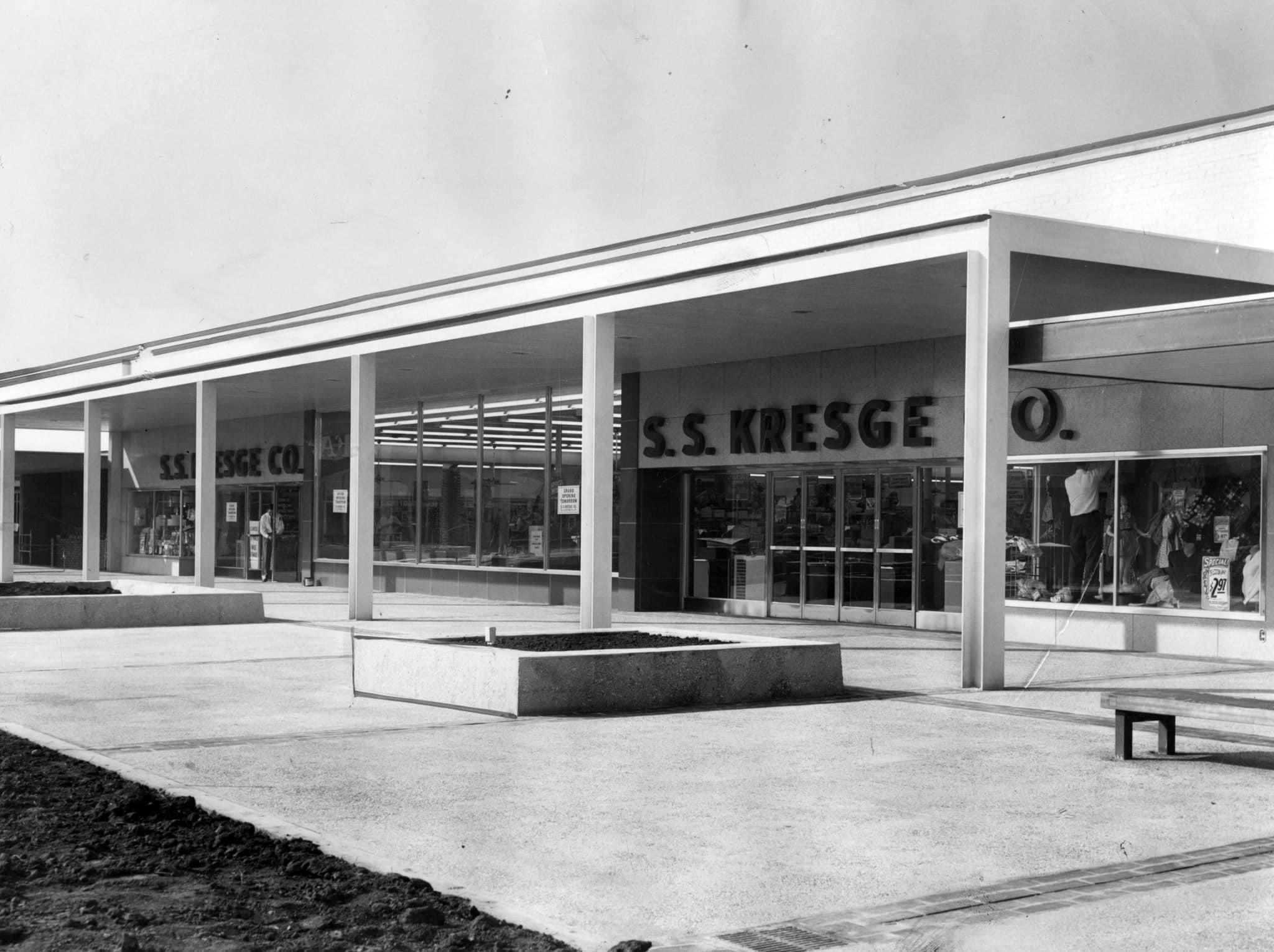Black and white photo of an S.S. Kresge Co. storefront with large windows and visible signage. The building features a covered walkway supported by columns and a mostly empty concrete plaza in front.
