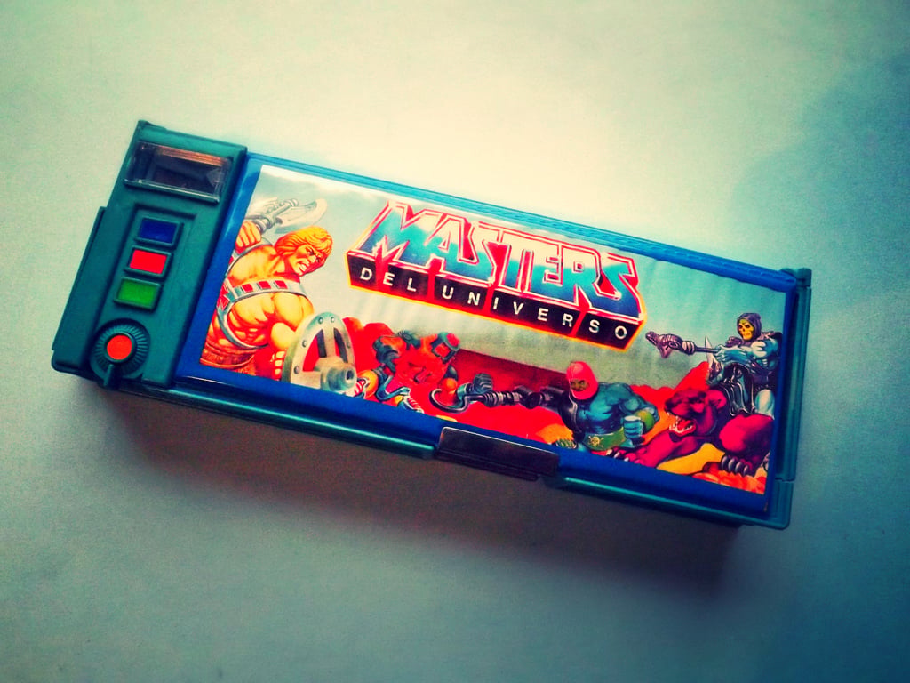 A colorful plastic pencil box features Masters del Universo characters in action poses, with bold red and blue text and a retro design.