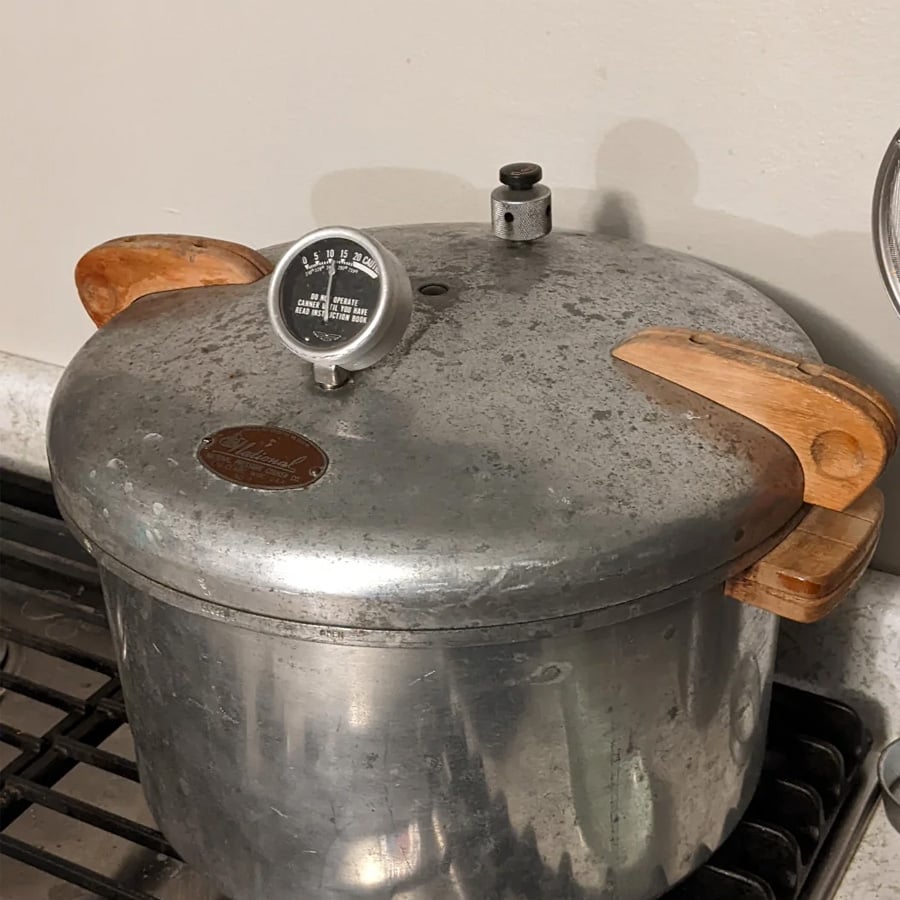 A vintage metal pressure cooker with wooden handles sits on a stove. A pressure gauge is attached to the lid, and the cooker shows signs of wear and age.