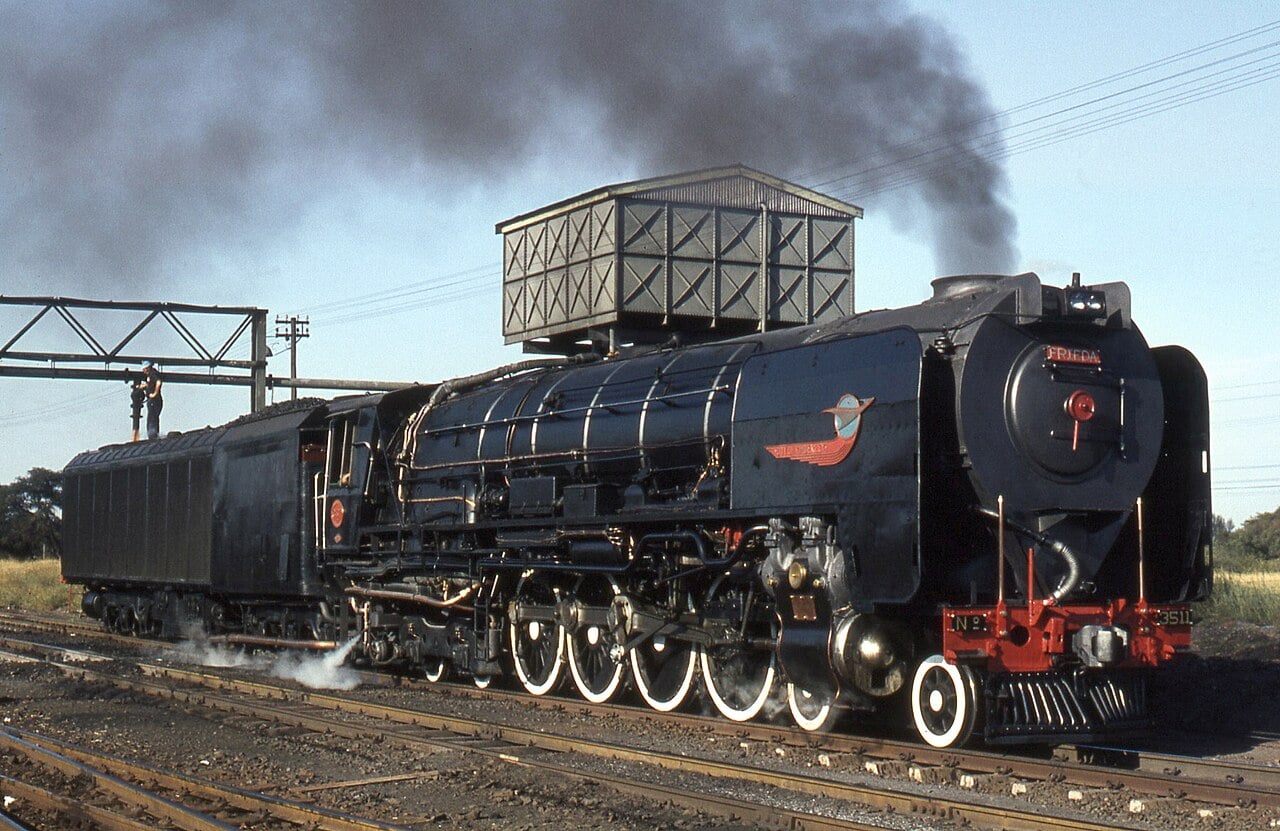 A black steam locomotive with white-rimmed wheels emits smoke, standing on railway tracks near a metal water tank and power lines, under a clear sky.