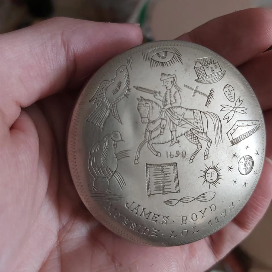 A hand holds a round engraved object showing a person on horseback, various symbols including birds, an eye, a star, a sun, a book, and the text “JAMES BOYD 1699 KOSSNET KOL 1777.”