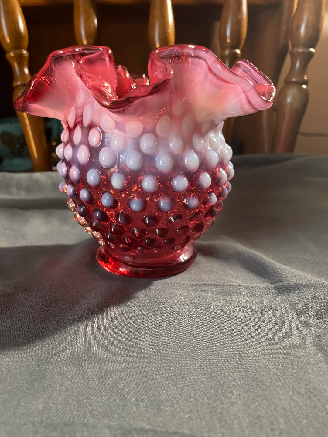A small, ruffled glass vase with a hobnail pattern in shades of pink and white sits on a gray cloth, with wooden chair backs in the background.