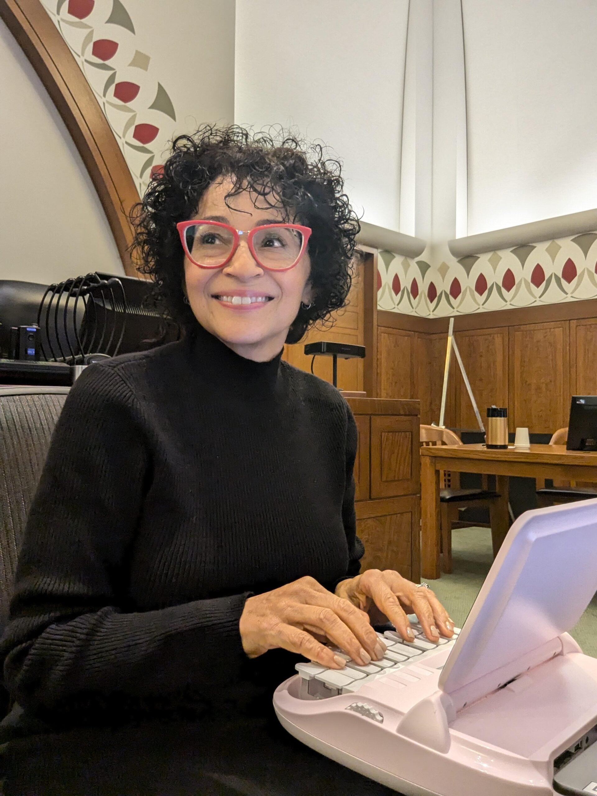 A smiling person with curly hair and pink glasses types on a white stenography machine in a wood-paneled room with patterned walls and a high ceiling.