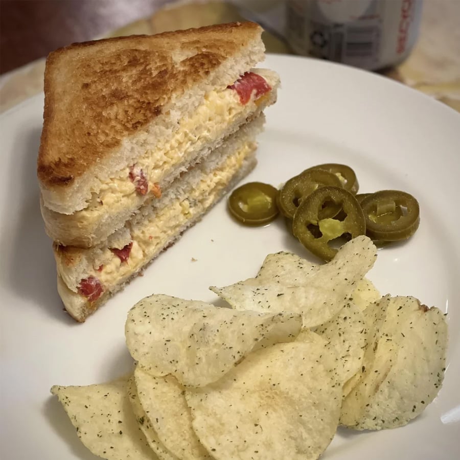 A plate with a pimento cheese sandwich cut in half, potato chips, and sliced pickled jalapeños. A drink can is partially visible in the background.