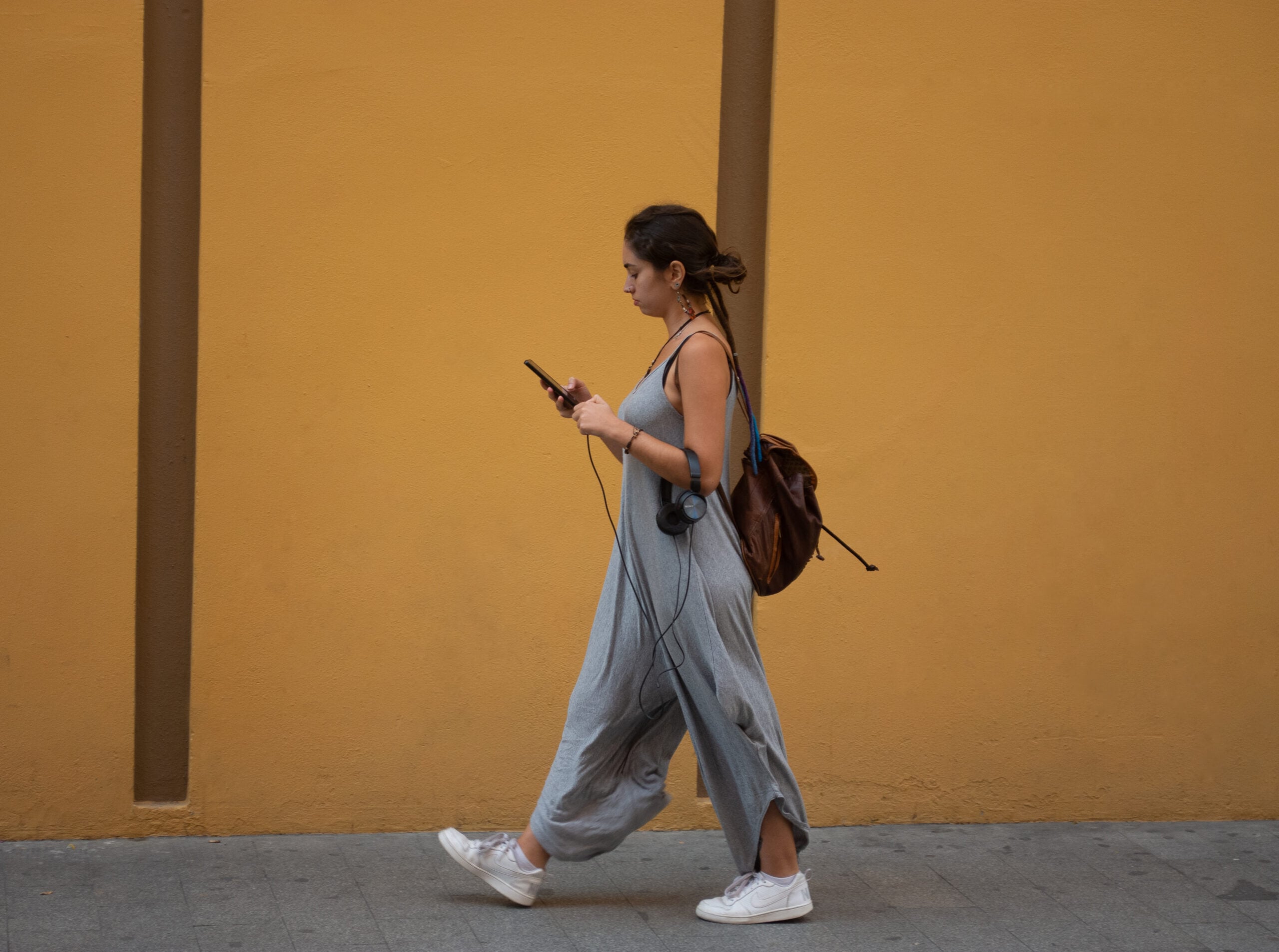 A woman in a gray jumpsuit walks past a yellow wall, holding her phone and headphones, with a brown backpack on her back.