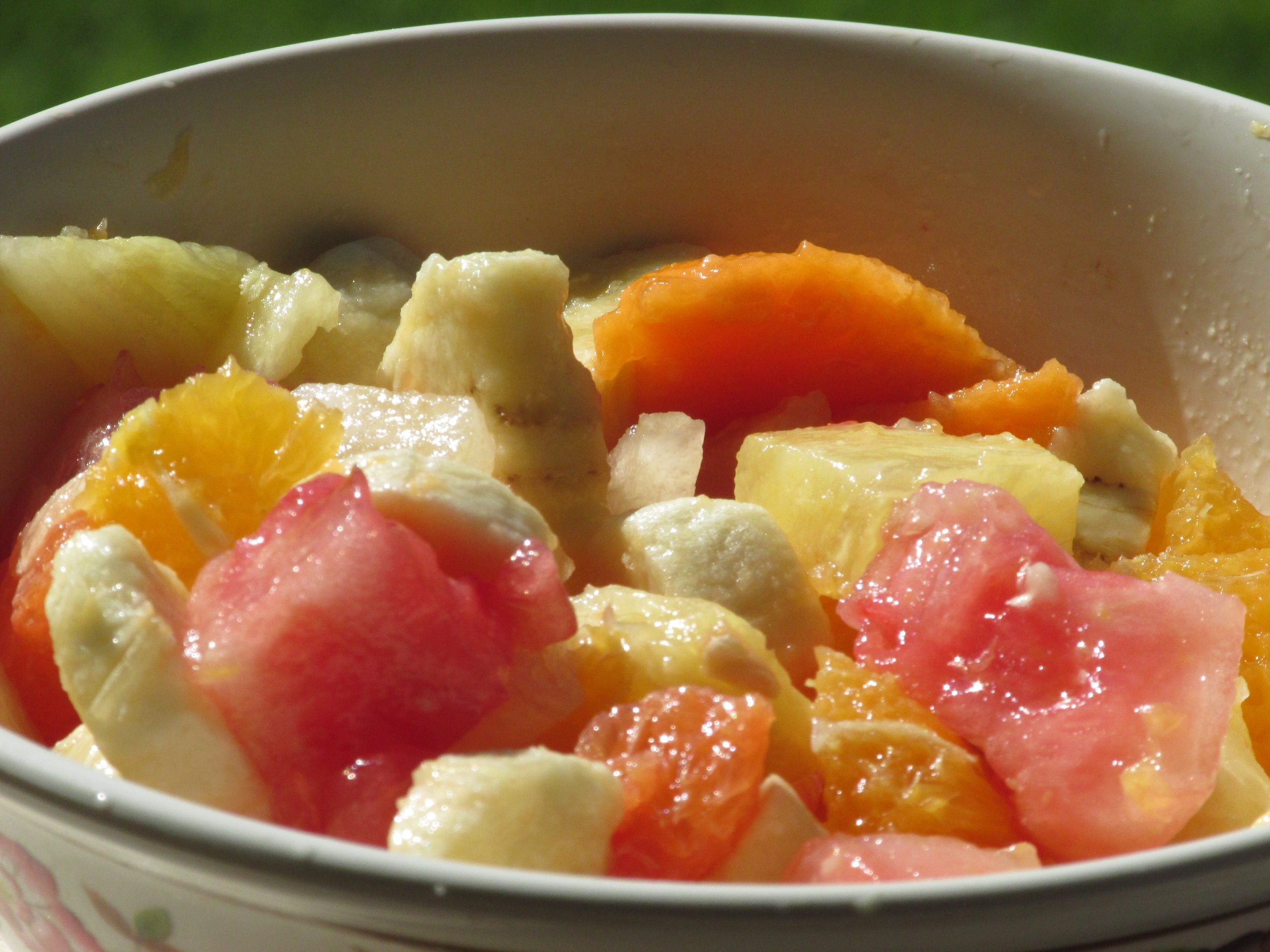 A close-up of a bowl filled with a fresh fruit salad, including pieces of banana, orange, watermelon, and papaya, with sunlight shining on the fruit.