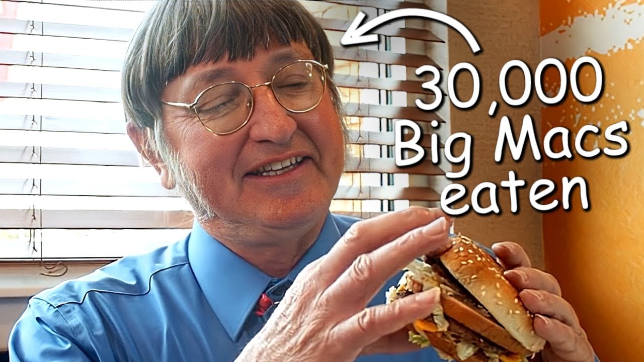 A smiling man in glasses and a blue shirt holds a Big Mac burger. Text on the image reads “30,000 Big Macs eaten” with an arrow pointing to him. Blinds and an orange wall are in the background.