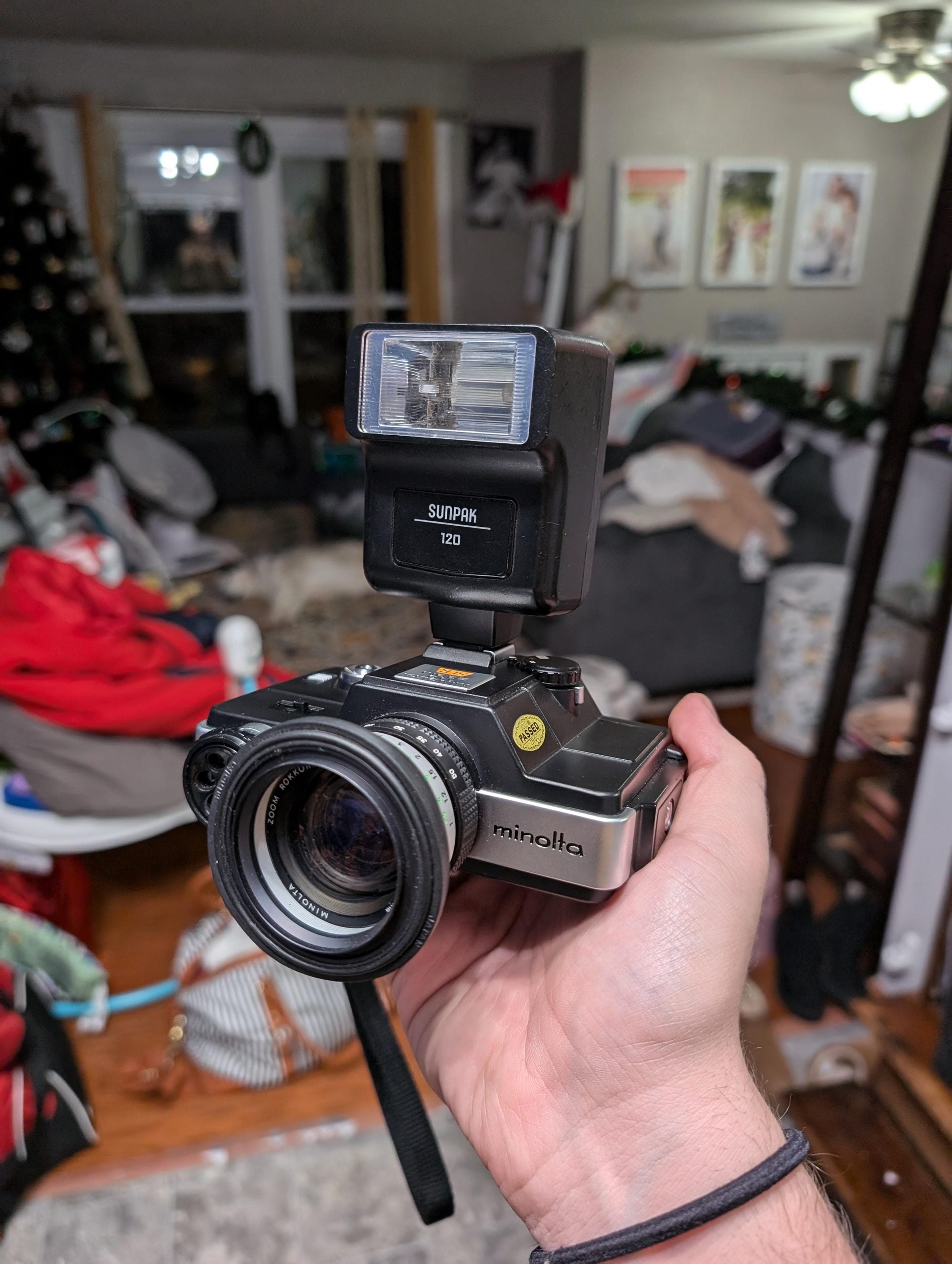 A hand holds a vintage Minolta camera with a Sunpak 120 flash attached. The background shows a cluttered living room with various items, a couch, and framed pictures on the wall.