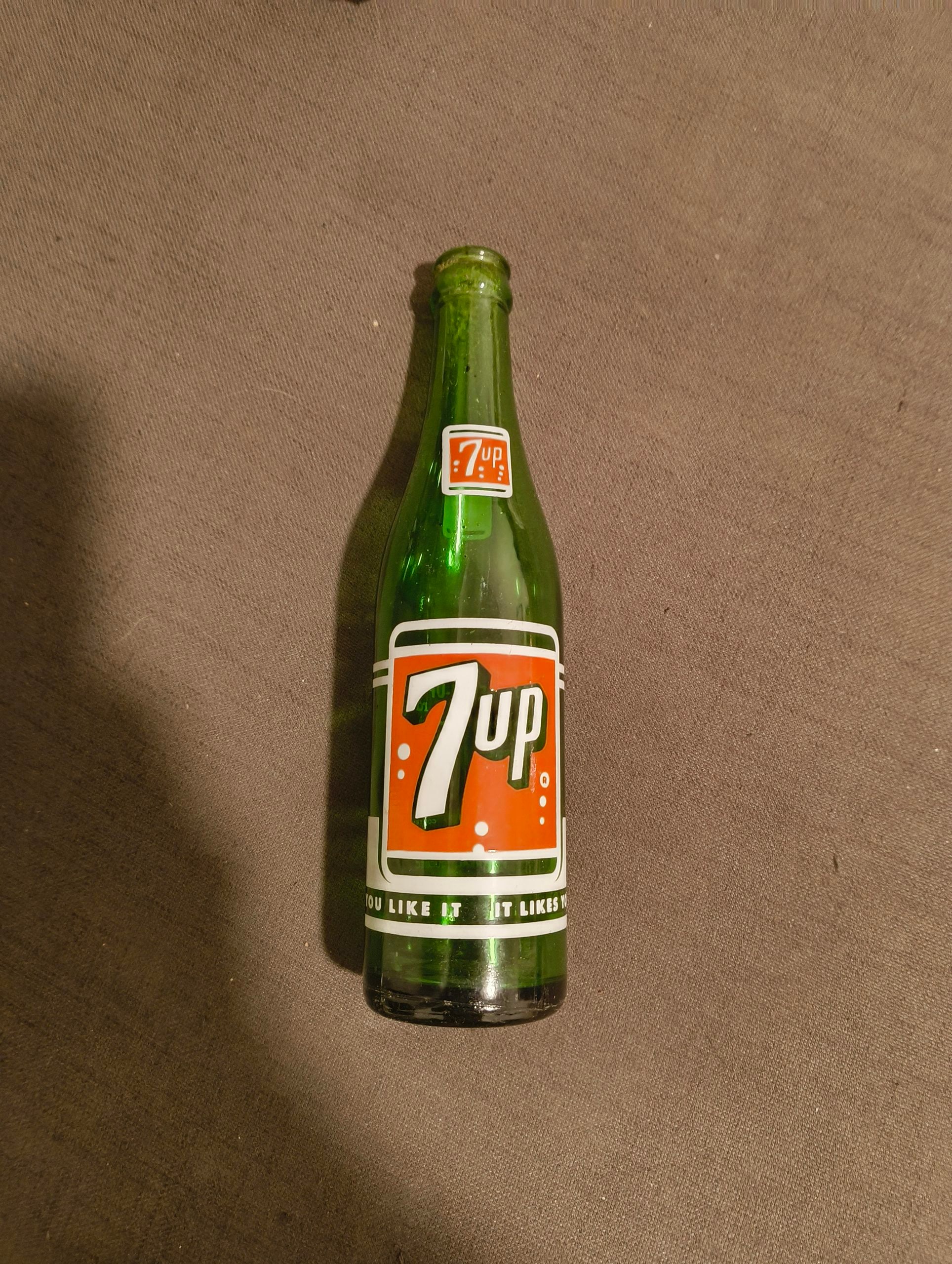 A vintage green glass 7Up bottle with a red, white, and black label lies on a brown fabric surface. The label features the 7Up logo and text near the bottom of the bottle.