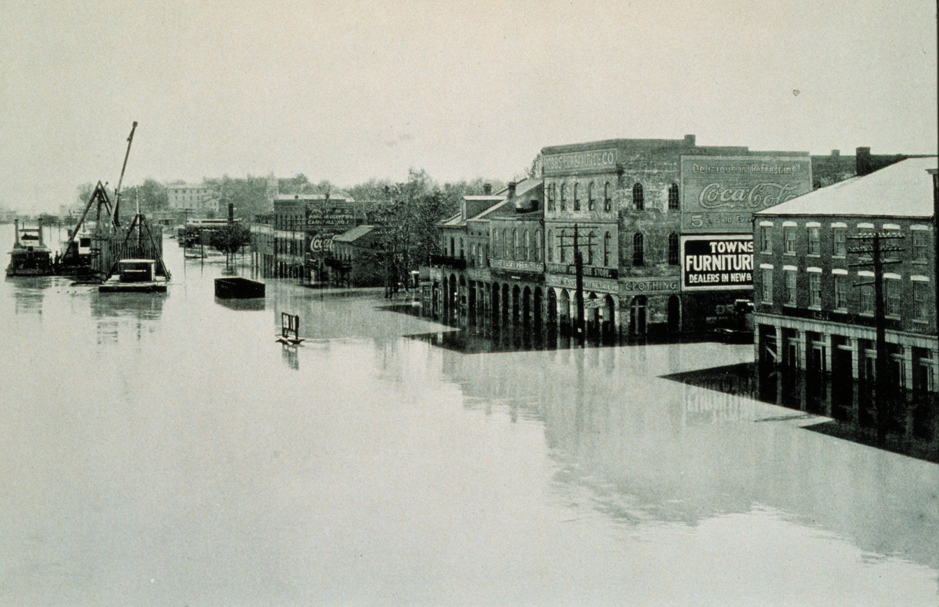 Historic black-and-white photo of a flooded town street; water covers the road up to the windows of brick buildings, with signs for furniture and Coca-Cola visible. Boats and cranes are seen on the left.