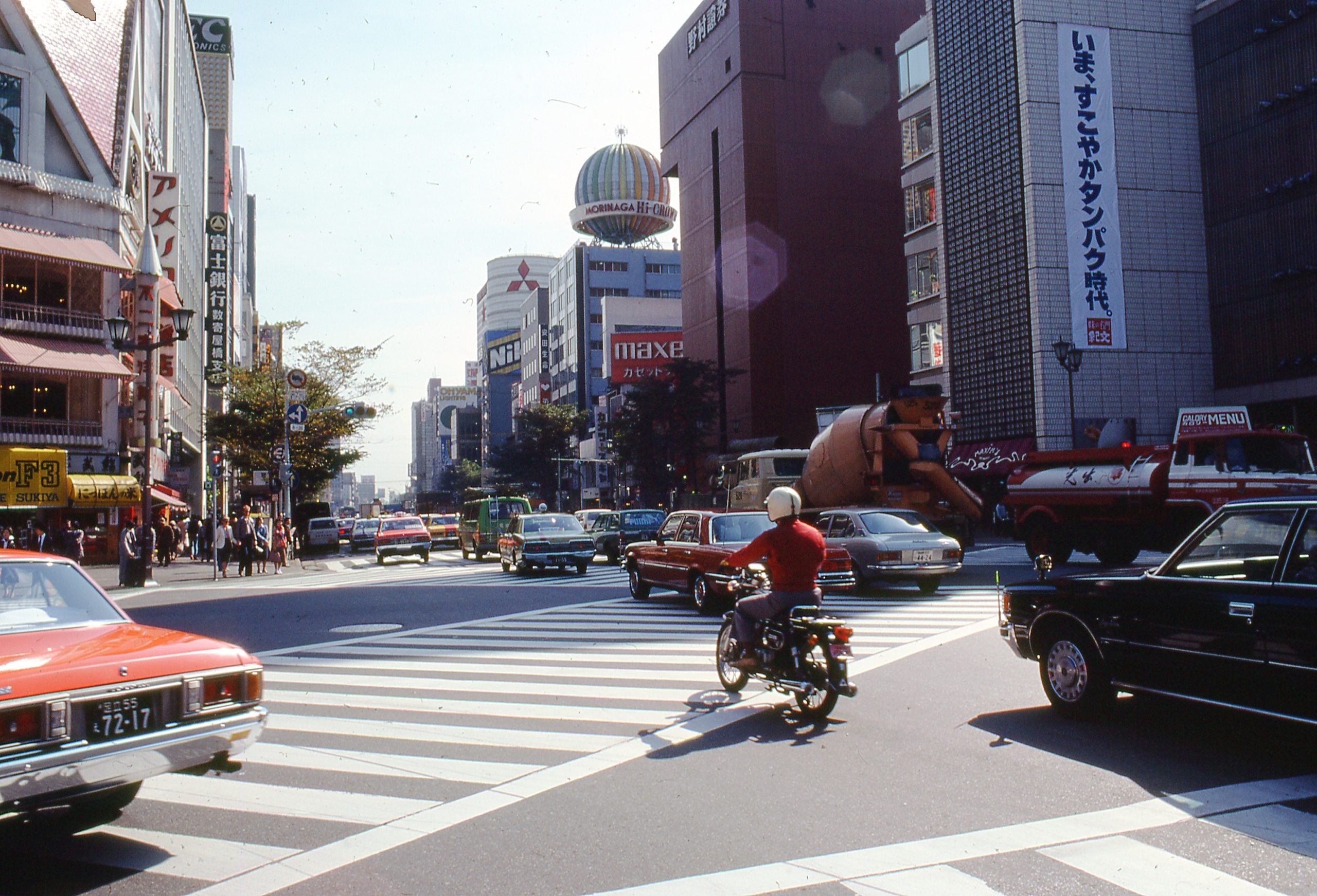 A busy city street in Japan with cars and a motorcyclist at a crosswalk, surrounded by tall buildings, shops, and billboards under a clear sky.
