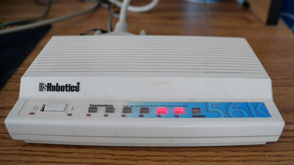 A white USRobotics 56K faxmodem with indicator lights and a power switch sits on a wooden surface. Several lights are illuminated on the front panel. Cables are plugged into the back.