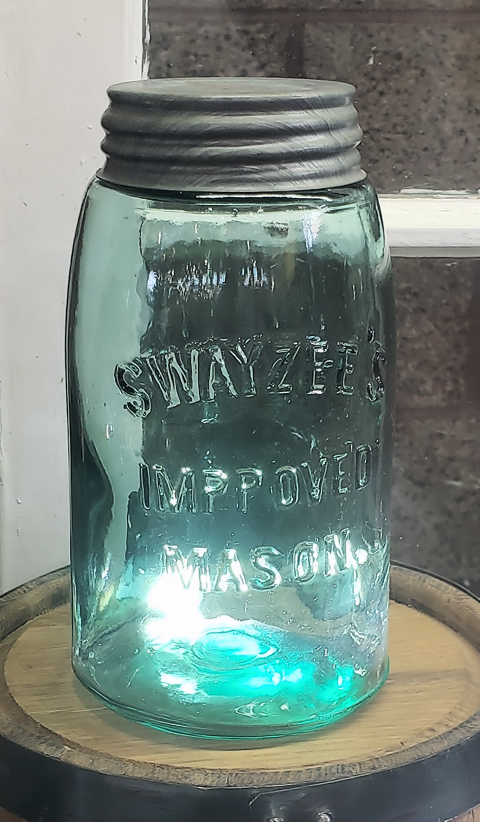 A vintage glass mason jar with a metal lid, embossed with the words “SWAYZEE’S IMPROVED MASON,” sits on a wooden surface against a window and brick background.