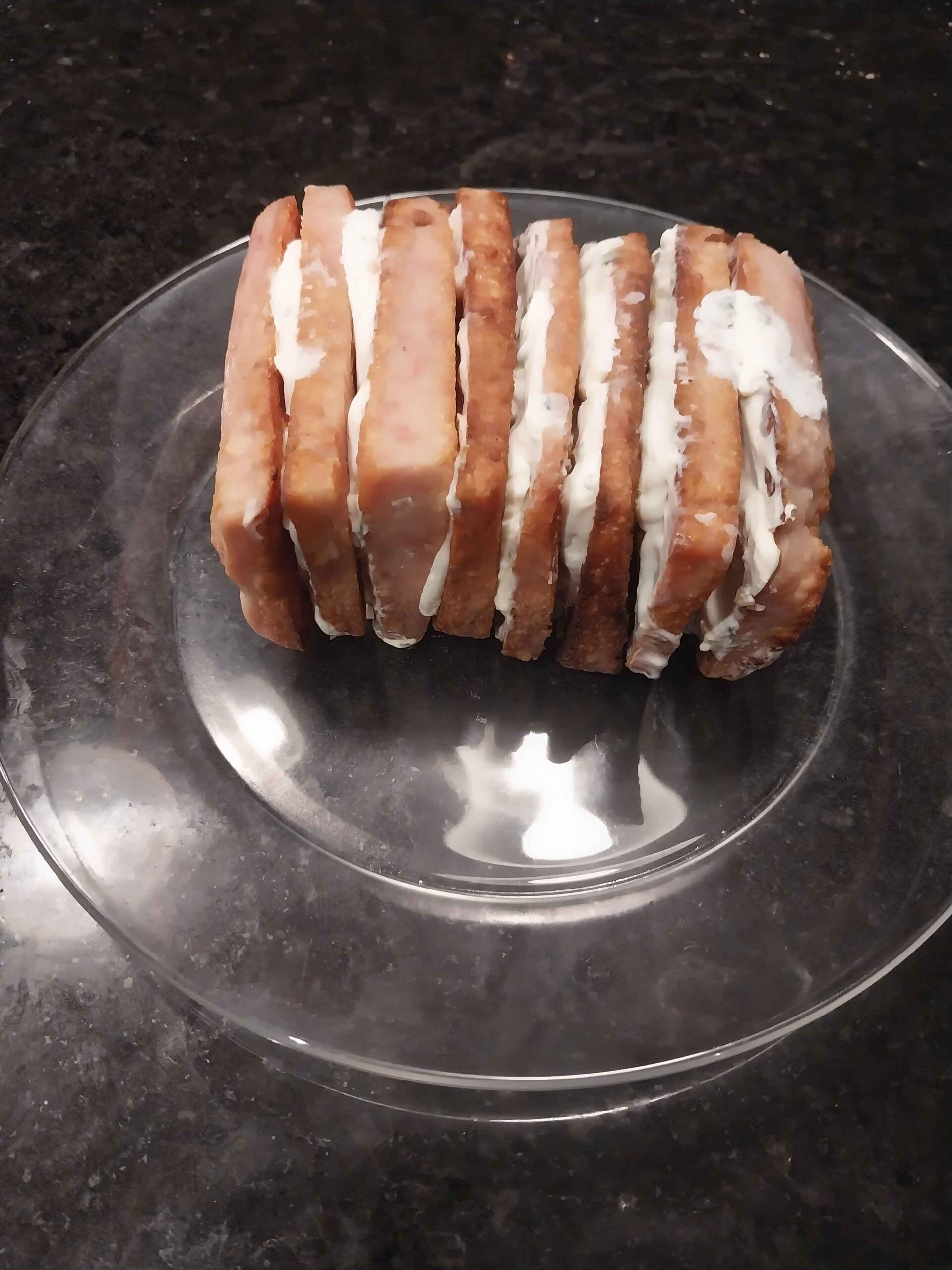 Sliced Spam pieces stacked upright on a clear plate, each slice filled with a layer of white spread, likely cream cheese, against a dark countertop background.