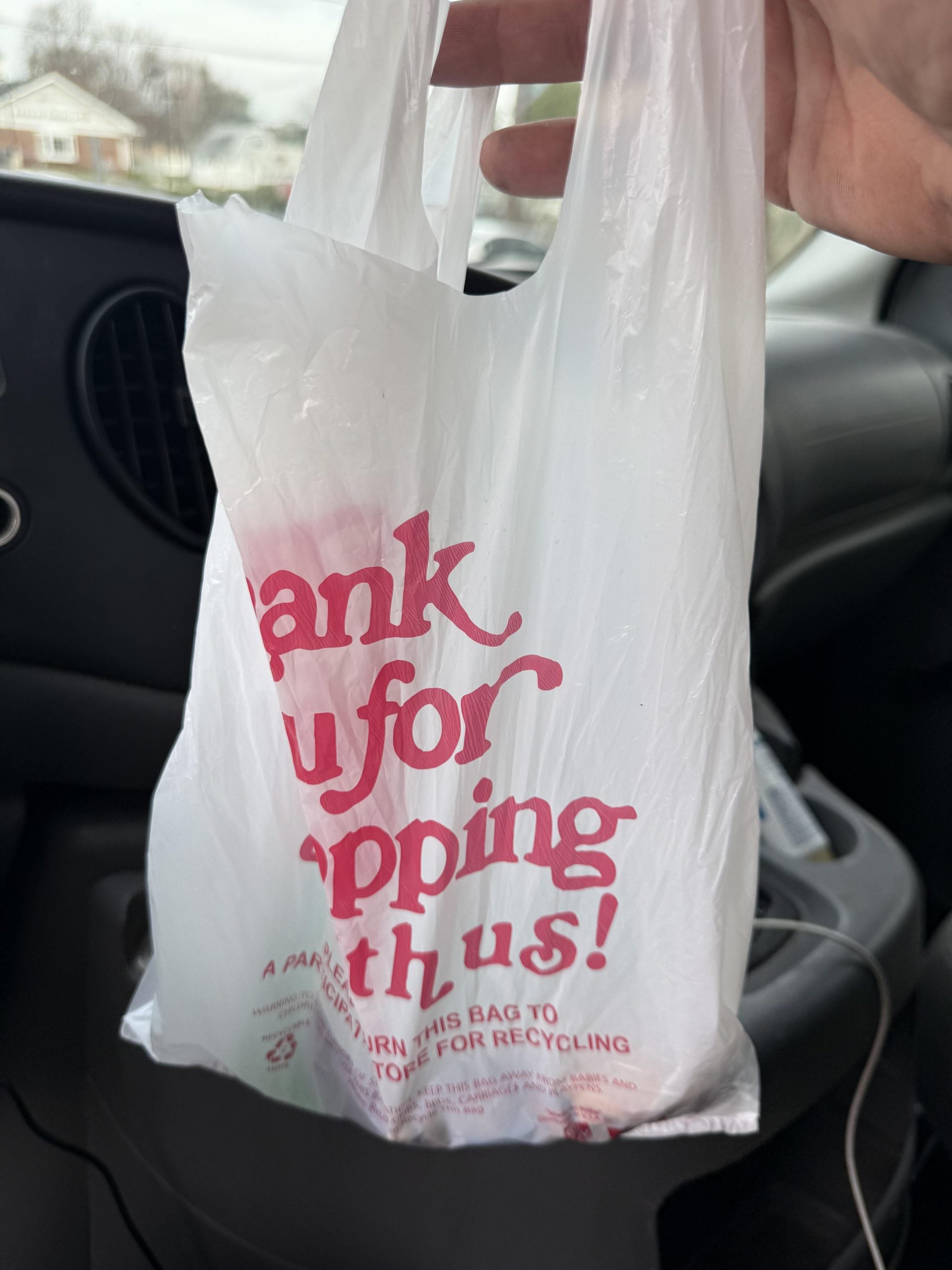 A hand holds a white plastic shopping bag with red text that reads “Thank you for shopping with us!” inside a car.
